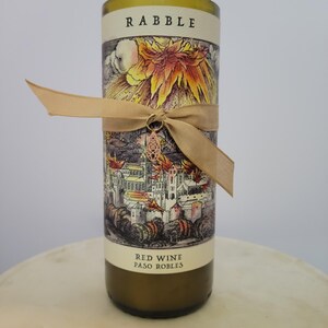 May include: A cylindrical wine bottle with a decorative label featuring a detailed illustration of a town and a fiery sun. The bottle is wrapped with a tan ribbon and a charm. The label reads "RABBLE" and "Red Wine Paso Robles."
