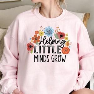 May include: A light pink sweatshirt with a floral design and the text "Helping Little Minds Grow" in black. The floral arrangement includes red, orange, blue, and yellow flowers. The sweatshirt has long sleeves.