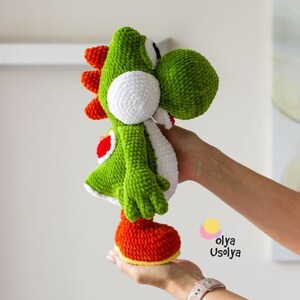 May include: A green and white crocheted plush toy featuring a large rounded snout, a white belly patch, and a red saddle on its back. The character has orange spikes along its spine and orange feet. The text Olya Usolya is printed in black.