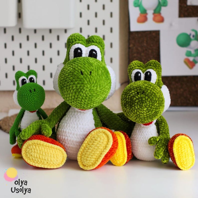 May include: Three crocheted plush figures of a green dinosaur character with white bellies, large white eyes with black pupils, and bright yellow and orange feet. The figures are arranged in a group, with one standing and two sitting.