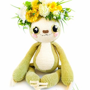 May include: A green crocheted sloth wearing a wreath of yellow and white flowers. The sloth has a white belly and a brown nose. The sloth is sitting with its legs crossed.