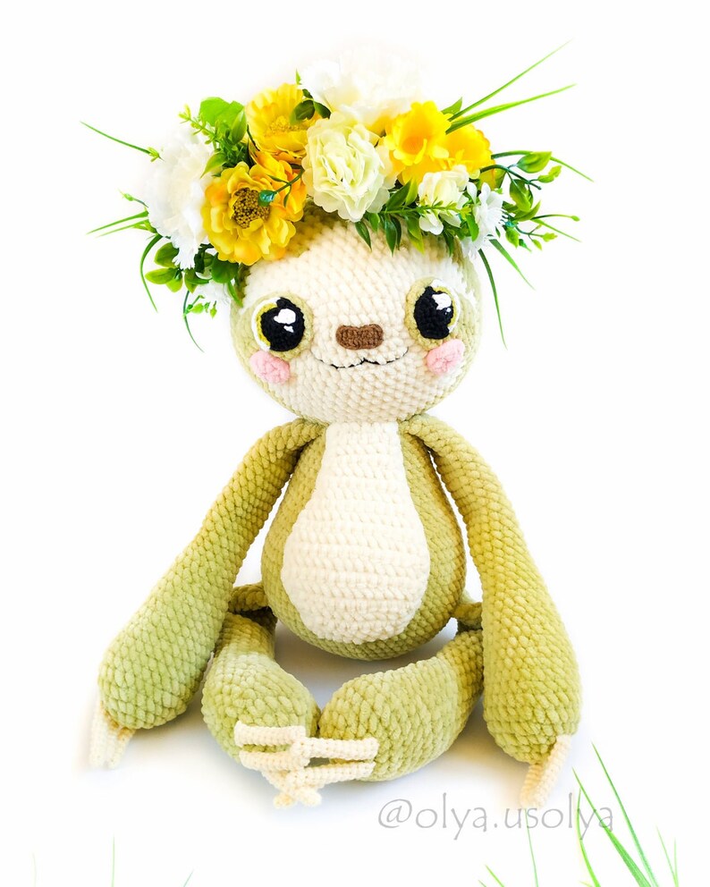 May include: A green crocheted sloth wearing a wreath of yellow and white flowers. The sloth has a white belly and a brown nose. The sloth is sitting with its legs crossed.