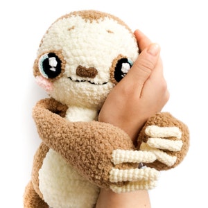 May include: A crocheted sloth plush toy with brown fur, white belly, and large black eyes. The sloth is being held by a hand.