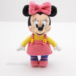 May include: A crocheted Minnie Mouse doll wearing a pink dress, yellow top, and black ears with a pink bow. The doll has a white face with black eyes and a black nose.