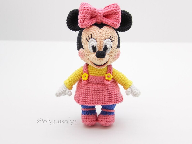 May include: A crocheted Minnie Mouse doll wearing a pink dress, yellow top, and black ears with a pink bow. The doll has a white face with black eyes and a black nose.