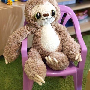 May include: A plush brown and white sloth toy with large eyes sits on a purple plastic chair. The chair is on a green carpet.