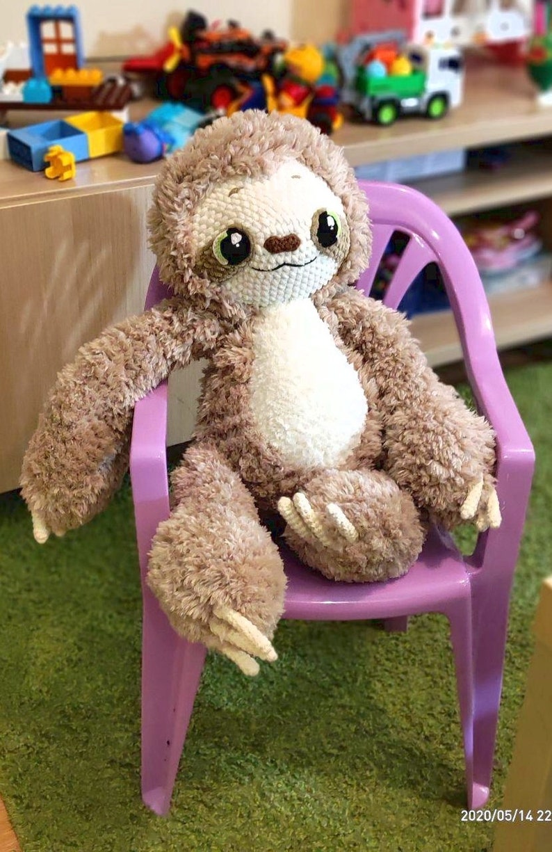 May include: A plush brown and white sloth toy with large eyes sits on a purple plastic chair. The chair is on a green carpet.