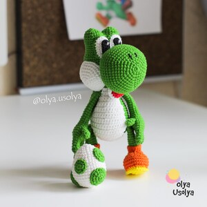 May include: A green and white crocheted dinosaur figure with large black and white eyes, a red mouth, and orange and yellow feet. The figure holds a white egg with green spots. The text @olya.usolya and olya usolya are visible on the image.