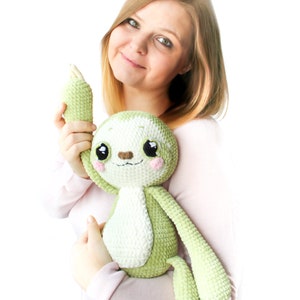 May include: A woman is holding a green and white crocheted sloth plush toy. The sloth has big eyes and a cute expression.