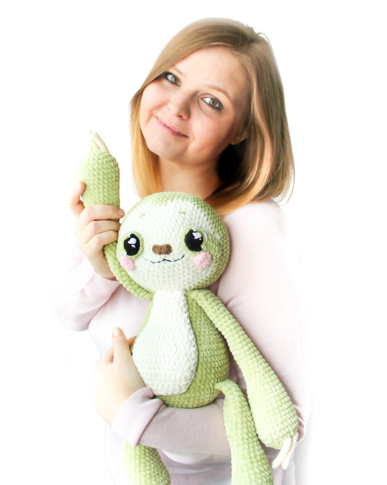 May include: A woman is holding a green and white crocheted sloth plush toy. The sloth has big eyes and a cute expression.