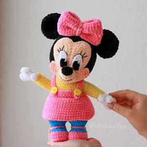 May include: A crocheted plush toy of a cartoon mouse wearing a pink dress, yellow shirt, and a pink bow. The toy has black ears, white gloves, and blue shoes with white stripes.