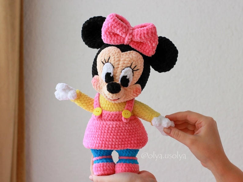 May include: A crocheted plush toy of a cartoon mouse wearing a pink dress, yellow shirt, and a pink bow. The toy has black ears, white gloves, and blue shoes with white stripes.