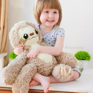 May include: A young girl wearing a white headband with bear ears holds a brown and white crocheted sloth plush toy. The girl is wearing a black and white polka dot shirt. The text '@olya.usolya' is visible in the image.
