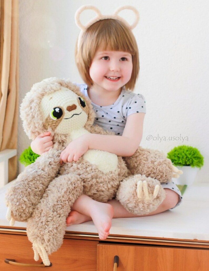 May include: A young girl wearing a white headband with bear ears holds a brown and white crocheted sloth plush toy. The girl is wearing a black and white polka dot shirt. The text '@olya.usolya' is visible in the image.