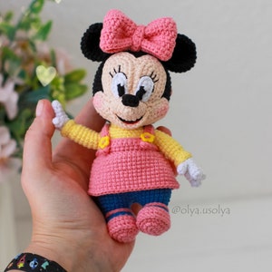 May include: A crocheted Minnie Mouse doll wearing a pink dress, yellow shirt, and blue trousers. She has a pink bow on her head and black ears. The doll is holding her hands up in the air. @olya.usolya