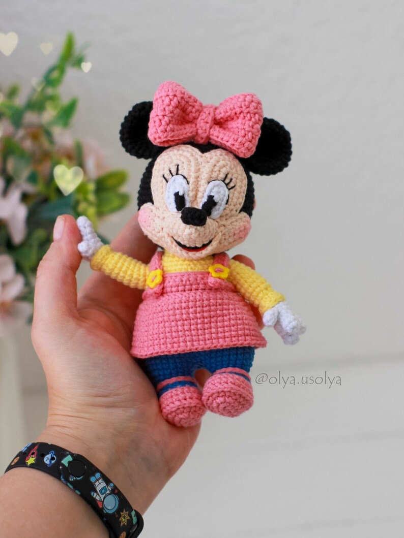 May include: A crocheted Minnie Mouse doll wearing a pink dress, yellow shirt, and blue trousers. She has a pink bow on her head and black ears. The doll is holding her hands up in the air. @olya.usolya