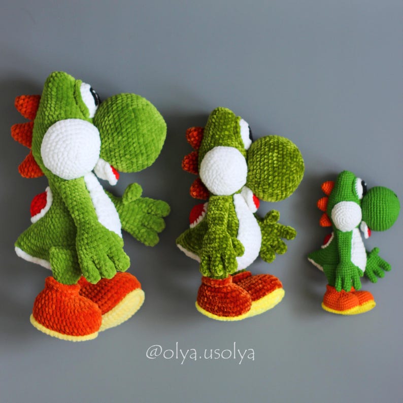 May include: Three handmade crochet plush toys of a green dinosaur character with white cheeks, orange spines, and orange shoes. The figures are arranged in descending order of size from left to right. The text @olya.usolya is visible at the bottom.