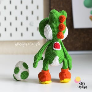 May include: A green crocheted dinosaur figure with white cheek patches, a red saddle, and orange spikes along its back. The figure wears orange shoes with yellow soles and stands next to a small white egg with green spots. Text reads @olya.usolya.