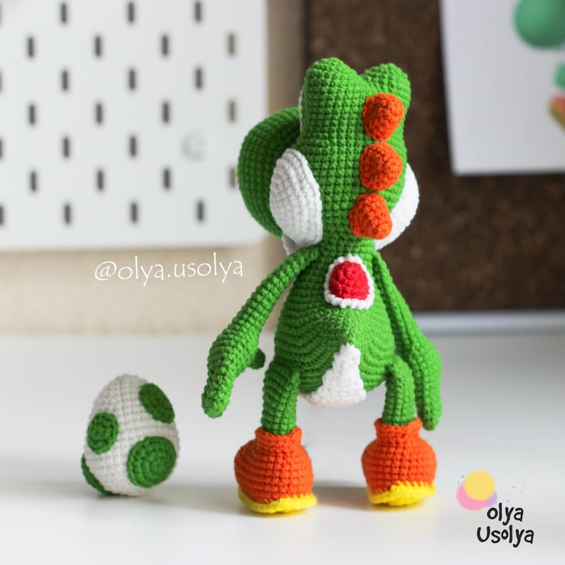 May include: A green crocheted dinosaur figure with white cheek patches, a red saddle, and orange spikes along its back. The figure wears orange shoes with yellow soles and stands next to a small white egg with green spots. Text reads @olya.usolya.