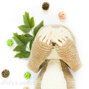 May include: A crocheted sloth toy with brown fur and a white belly. The sloth is covering its eyes with its paws. The toy is surrounded by small balls of yarn in various colors.