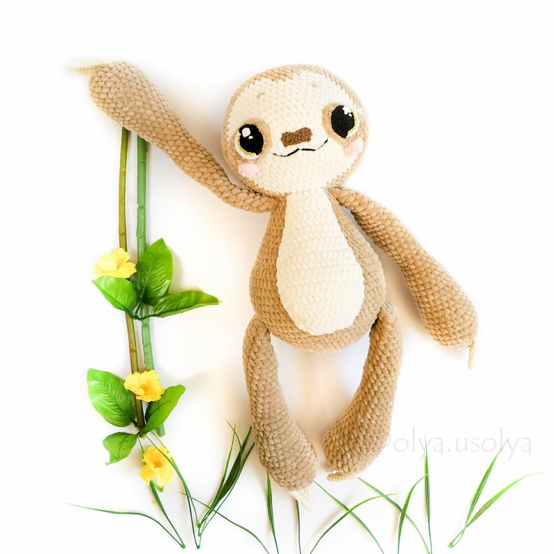 May include: A crocheted sloth toy with brown fur and a white belly. The sloth is hanging from a green vine with yellow flowers. The toy is on a white background.