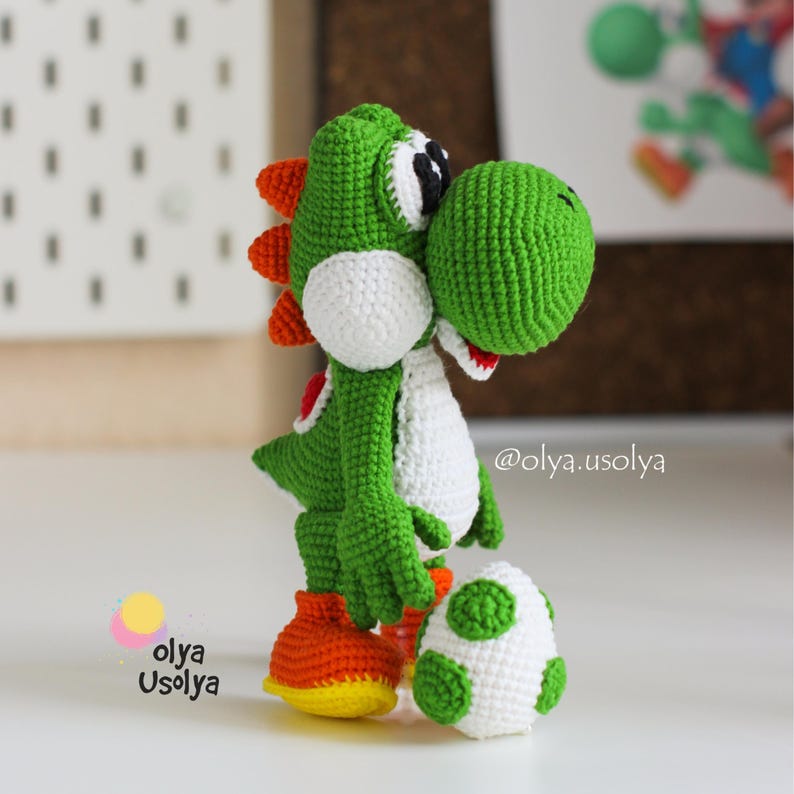 May include: A green and white crocheted Yoshi figure with orange spikes on its back and orange shoes. The figure stands next to a small white egg with green spots. The text @olya.usolya and Olya Usolya are visible in the lower left and right areas.