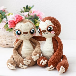 May include: Two crocheted sloth plushies, one brown and one cream, sitting together. The cream sloth has a pink bow on its head and both sloths have embroidered eyes and smiling faces.