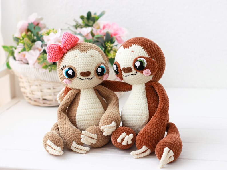 May include: Two crocheted sloth plushies, one brown and one cream, sitting together. The cream sloth has a pink bow on its head and both sloths have embroidered eyes and smiling faces.