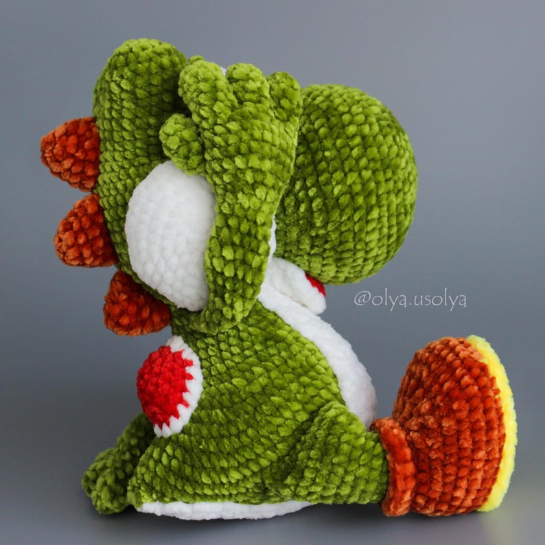 May include: A plush crochet toy shaped like a green dinosaur with a white belly, orange spikes along its back, and orange feet with yellow soles. The toy has a textured, soft appearance and features the text @olya.usolya printed on the side.