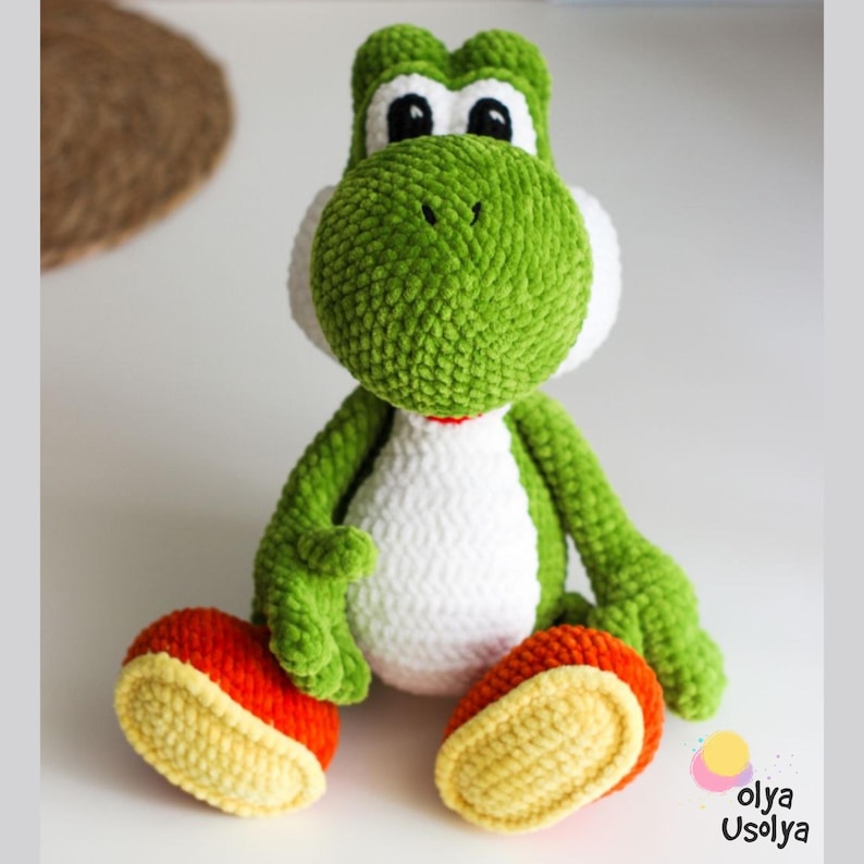 May include: A green and white crocheted plush toy resembling a dinosaur character with large black eyes, a rounded snout, and orange and yellow feet. The figure has a soft, textured yarn appearance and is sitting in an upright position.