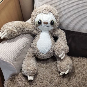 May include: A crocheted sloth plush toy with brown fur and a white belly. The sloth is sitting on a brown fuzzy surface.