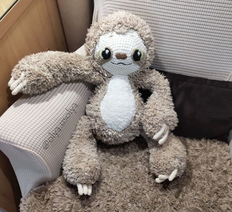 May include: A crocheted sloth plush toy with brown fur and a white belly. The sloth is sitting on a brown fuzzy surface.