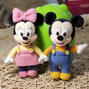 May include: Two crocheted stuffed toys, Mickey Mouse and Minnie Mouse, are sitting on a woven surface. Mickey Mouse is wearing blue overalls and a yellow shirt. Minnie Mouse is wearing a pink dress and a yellow shirt with a pink bow.