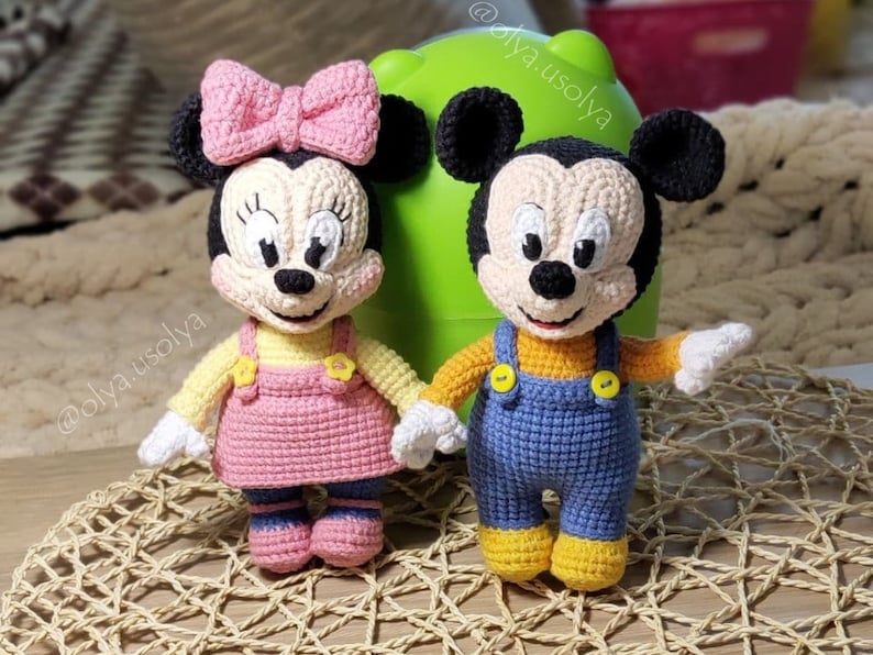 May include: Two crocheted stuffed toys, Mickey Mouse and Minnie Mouse, are sitting on a woven surface. Mickey Mouse is wearing blue overalls and a yellow shirt. Minnie Mouse is wearing a pink dress and a yellow shirt with a pink bow.