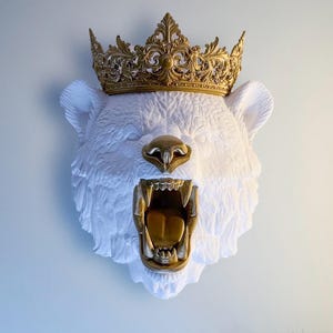May include: A white bear head wall sculpture with a gold crown and gold accents on the nose and mouth. The bear's mouth is open, revealing gold teeth and a tongue. The crown is ornate and detailed.