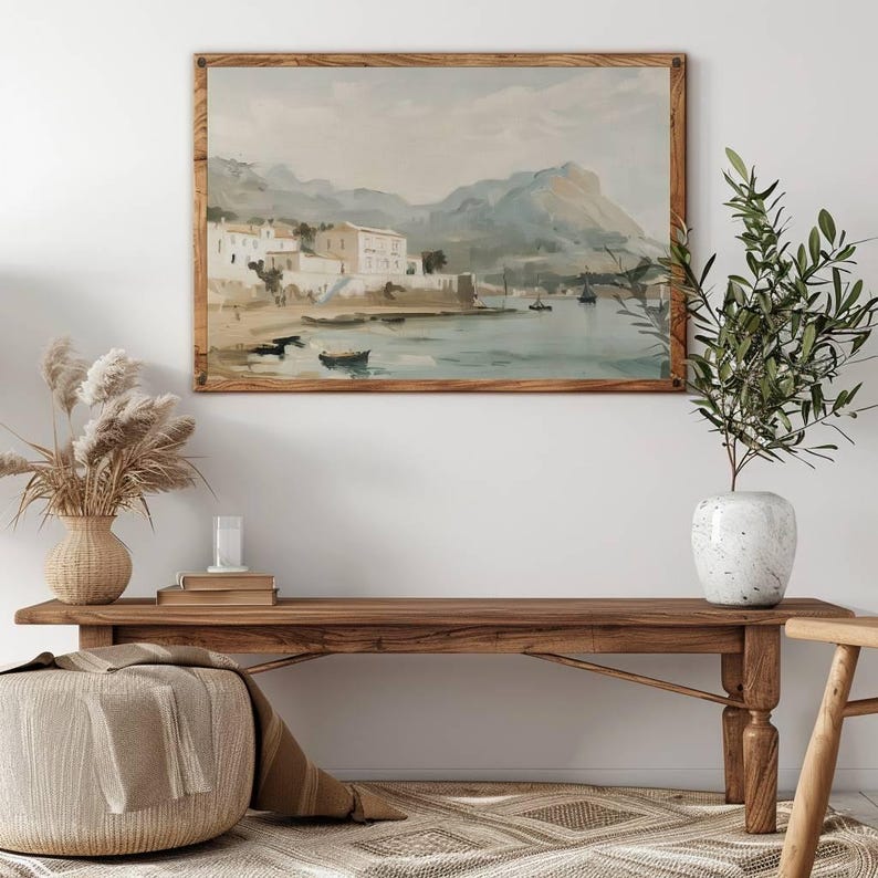 May include: A framed watercolor painting depicting a coastal town with white buildings, a harbor, and boats. The painting is set against a backdrop of rolling hills and a blue sky.