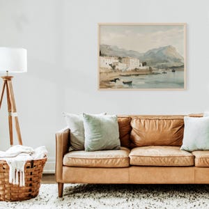 May include: A watercolor painting of a coastal town with white buildings and a mountain range in the background. The painting is framed in a light wood frame and hangs on a white wall above a brown leather couch.