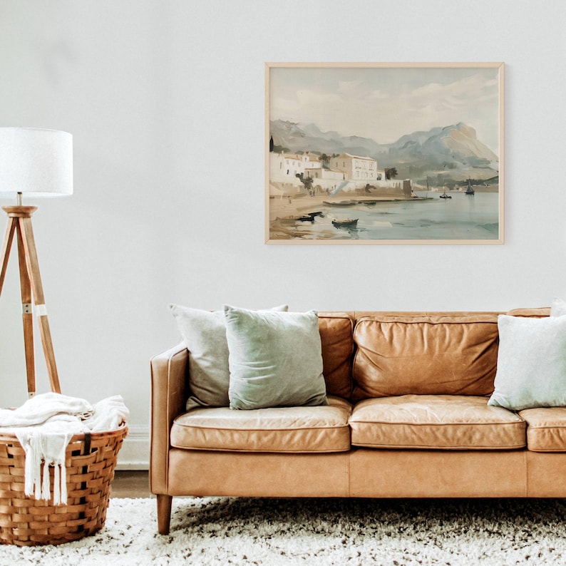 May include: A watercolor painting of a coastal town with white buildings and a mountain range in the background. The painting is framed in a light wood frame and hangs on a white wall above a brown leather couch.