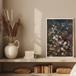 May include: A framed print of a floral arrangement featuring a variety of white, pink, and purple flowers on a dark background. The print is hanging on a beige wall above a shelf with books and a vase of dried grasses.