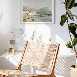 May include: A framed watercolor painting of a coastal town with buildings and mountains in the background. The painting is hanging on a white wall above a wicker chair.