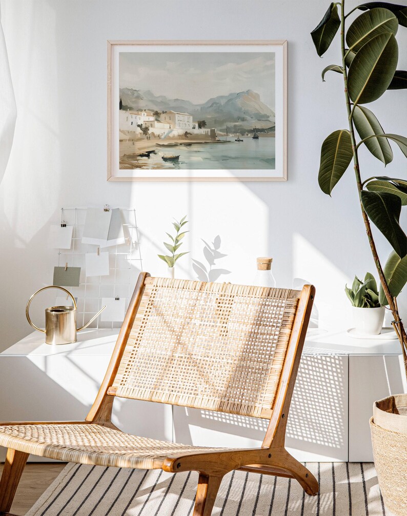 May include: A framed watercolor painting of a coastal town with buildings and mountains in the background. The painting is hanging on a white wall above a wicker chair.