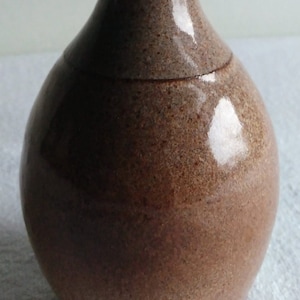 May include: A handcrafted ceramic vase with a slender neck and rounded body. The vase features a speckled brown and tan glaze with a blue-gray top. The vase is approximately 8 inches tall.