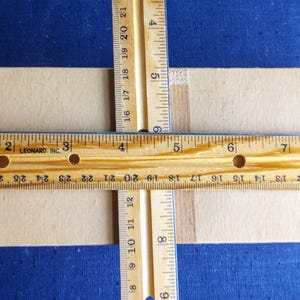 Puede incluir: Primer plano de una caja de cart&oacute;n beige con dos reglas de madera que se cruzan en &aacute;ngulo recto. Una regla mide en pulgadas, la otra en pulgadas y cent&iacute;metros. El fondo es azul.