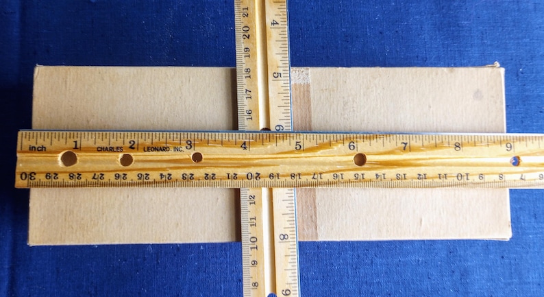 Puede incluir: Primer plano de una caja de cart&oacute;n beige con dos reglas de madera que se cruzan en &aacute;ngulo recto. Una regla mide en pulgadas, la otra en pulgadas y cent&iacute;metros. El fondo es azul.