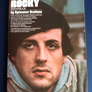 Puede incluir: El Scrapbook oficial de Rocky por Sylvester Stallone. La portada presenta un retrato de Sylvester Stallone y texto que describe la historia de la película, los premios y las nominaciones. El libro es una condensación fotográfica de la película ganadora del Oscar.