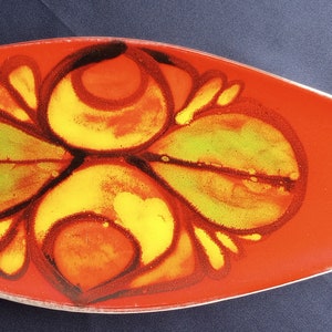 Puede incluir: Una fuente larga de cerámica naranja con un diseño floral en amarillo, verde y marrón. La fuente tiene un extremo puntiagudo y un borde blanco. El diseño floral está centrado en la fuente.