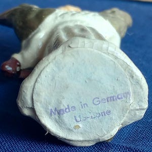 Puede incluir: Una peque&ntilde;a figura vintage de un perro, vista desde abajo. El perro es principalmente blanco con marcas marrones y negras. La base tiene una etiqueta de papel que dice "Made in Germany Us-zone".