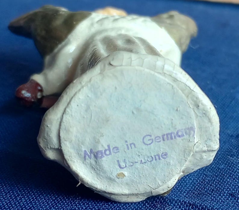 Puede incluir: Una peque&ntilde;a figura vintage de un perro, vista desde abajo. El perro es principalmente blanco con marcas marrones y negras. La base tiene una etiqueta de papel que dice "Made in Germany Us-zone".