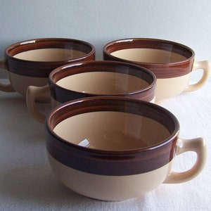 Puede incluir: Cuatro cuencos de sopa de cerámica con asas. Cada cuenco tiene un interior beige claro y una banda marrón oscuro alrededor del borde y la base. El exterior es beige claro con una franja marrón oscuro. Los cuencos están dispuestos sobre una superficie blanca.