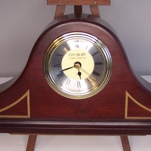Puede incluir: Un reloj de chimenea de madera con un borde dorado y una esfera blanca. La esfera del reloj tiene marcadores de hora en números romanos negros y agujas negras. La esfera del reloj también tiene el texto "Danbury Clock Company" impreso en ella.